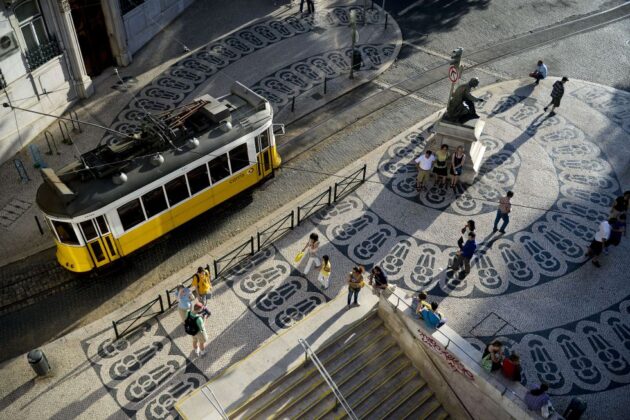 Tranvía amarillo circulando por una plaza de Lisboa con pisos de mosaicos portugueses y turistas caminando alrededor.