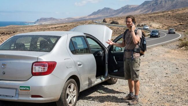 Auto de alquiler detenido en la ruta con el capó abierto y un viajero pidiendo ayuda por una avería durante el viaje.