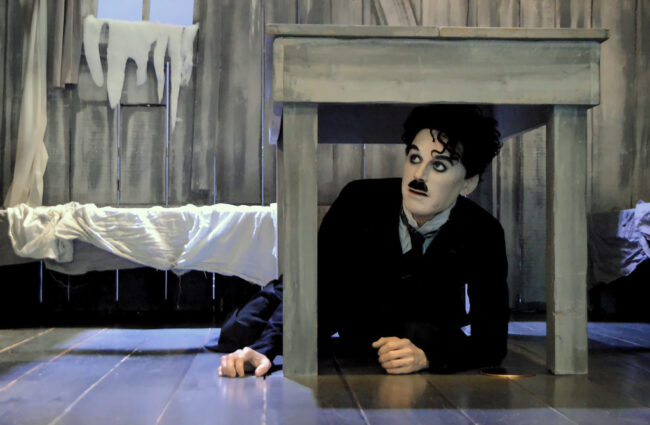 Escena de Charlie Chaplin en una cabaña, escondiéndose bajo una mesa