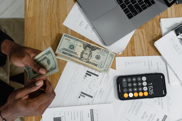 Presupuesto de viaje en dólares Persona contando billetes de dólar con laptop y calculadora sobre mesa de trabajo