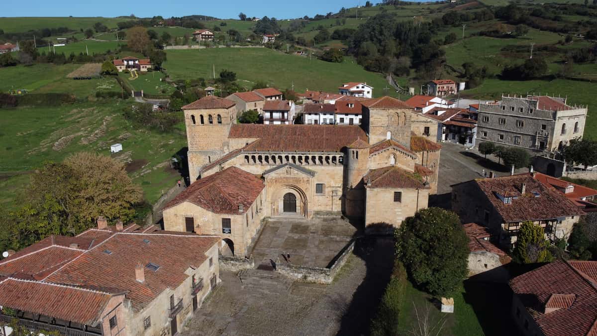 Vista aérea de la Colegiata de Santa Juliana en Santillana del Mar, rodeada de casas con techos rojos y prados verdes.