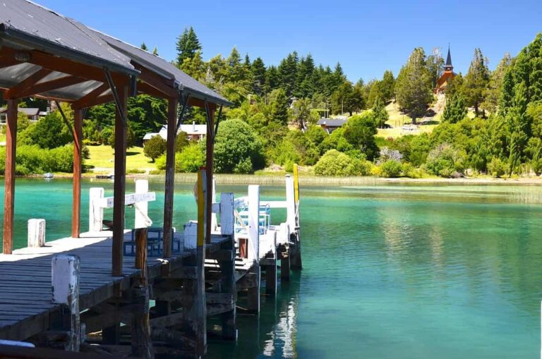Muelle de madera sobre lago de aguas turquesas en Bariloche, rodeado de bosque y cabañas.