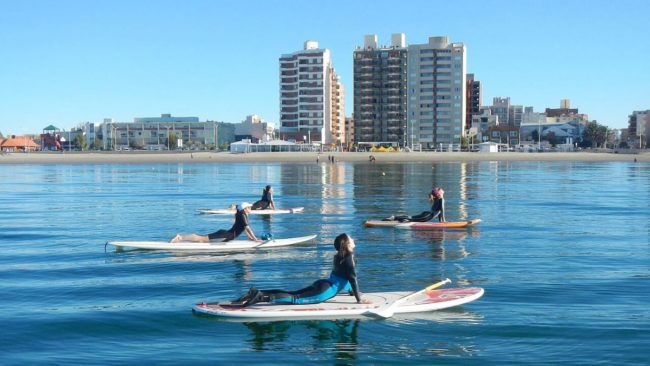 Paddle surf en las playas de Puerto Madryn Grupo de personas practicando paddle surf en las tranquilas aguas del Golfo Nuevo, con vista a los edificios del centro de Puerto Madryn.