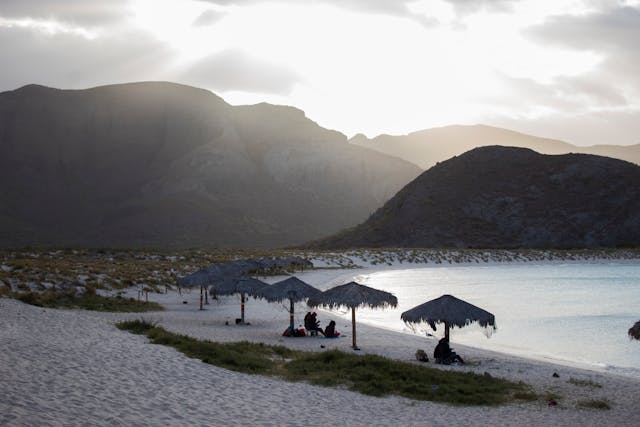 Palapas sobre la arena en una playa desierta rodeada de montañas al atardecer.