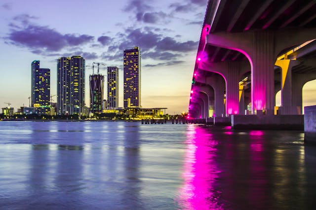 Vista nocturna de rascacielos en Miami y puente iluminado con luces rosas sobre el agua.