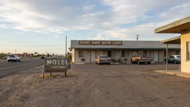 Qué es un motel y en qué se diferencia de un hotel (guía para viajeros) Motel de carretera en Estados Unidos con estacionamiento frente a las habitaciones y cartel de vacancy.