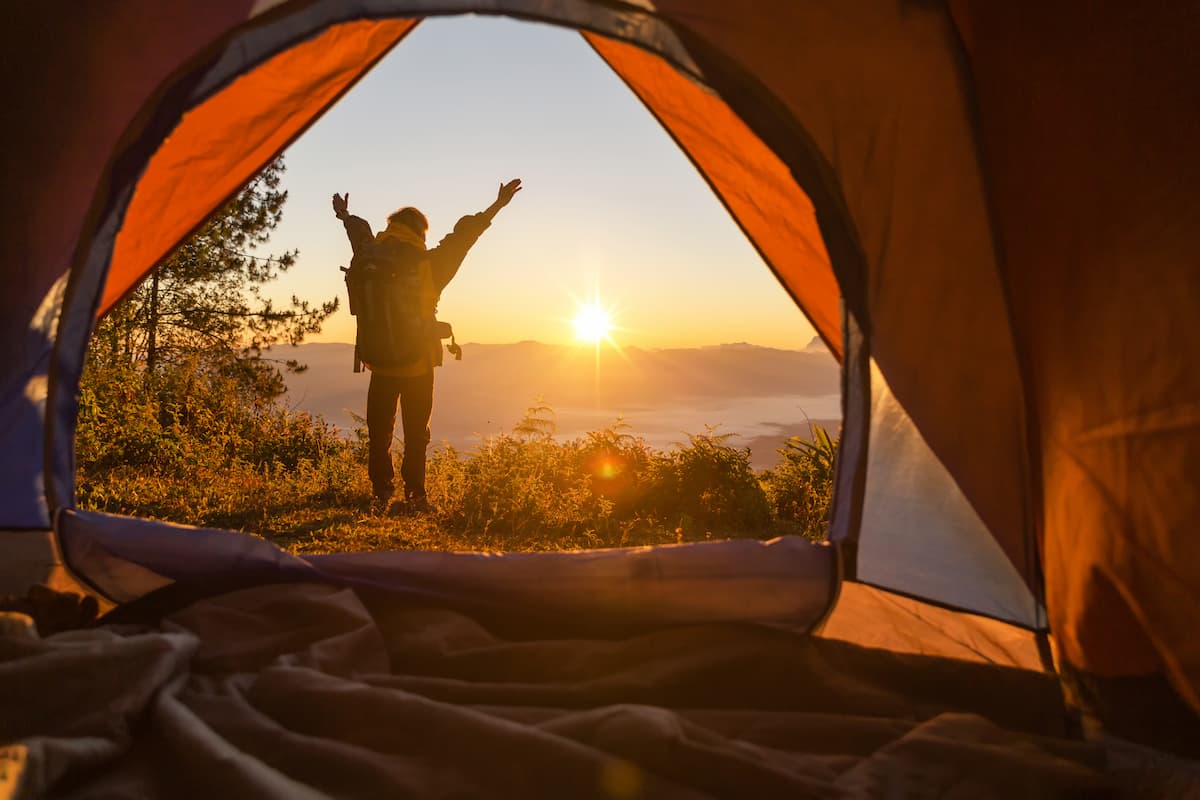 Excursionista se para frente a la tienda color naranja de campamento con la mochila mirando las montañas