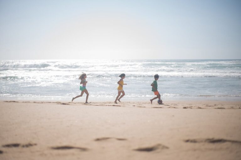 Niños corriendo y jugando al fútbol en la arena de una playa soleada.