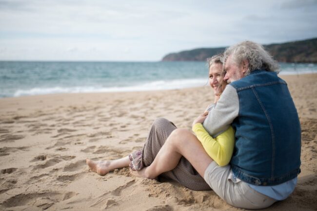 Pareja de adultos mayores abrazándose y sonriendo sentados en la playa.