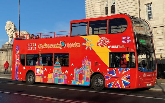 Autobús turístico City Sightseeing London frente al monumento del león. Autobús de dos pisos de City Sightseeing London en color rojo, con diseños turísticos coloridos y logotipo de Julià Travel.