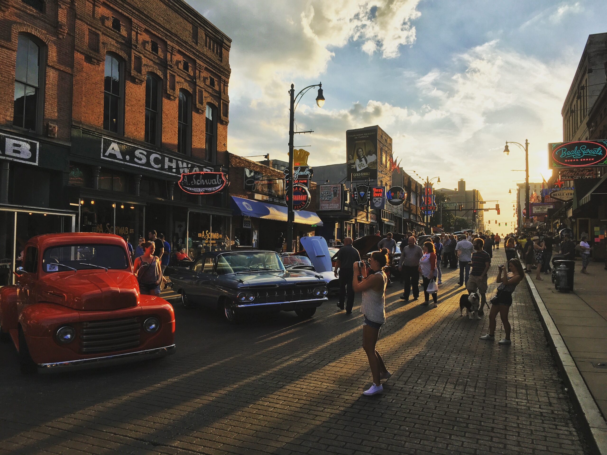 Personas caminando por Beale Street al atardecer en Memphis, rodeadas de autos clásicos y letreros de neón.