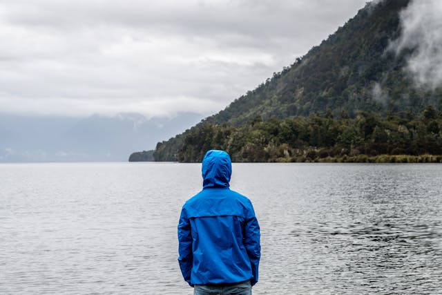 Exploración en solitario junto al lago, rodeado de montañas y niebla. Persona con campera azul observando un lago rodeado de montañas en un día nublado.