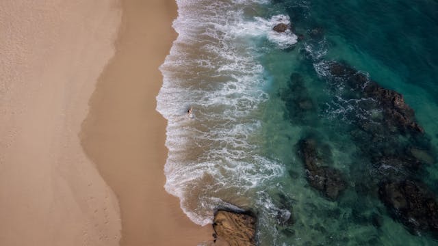 Vista aérea de una playa mexicana con aguas claras y formaciones rocosas. Persona en la orilla de una playa de arena dorada con agua turquesa y rocas sumergidas vistas desde un dron.