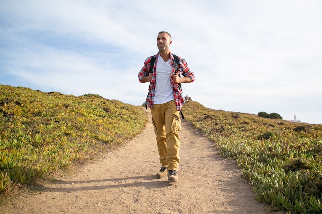 Hombre haciendo senderismo por un camino natural bajo el cielo despejado. Hombre caminando por un sendero de tierra rodeado de vegetación, con mochila y ropa de senderismo.
