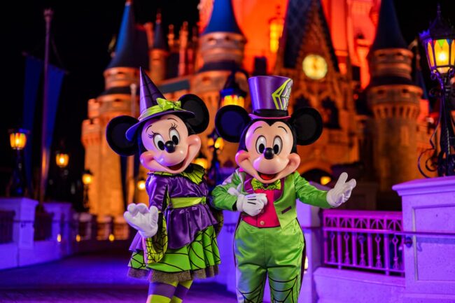 Mickey y Minnie disfrazados de Halloween posan frente al castillo de Disney iluminado de noche.