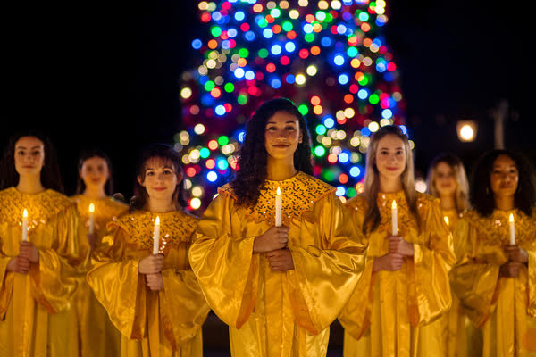 Coro navideño con túnicas doradas cantando con velas frente a árbol iluminado