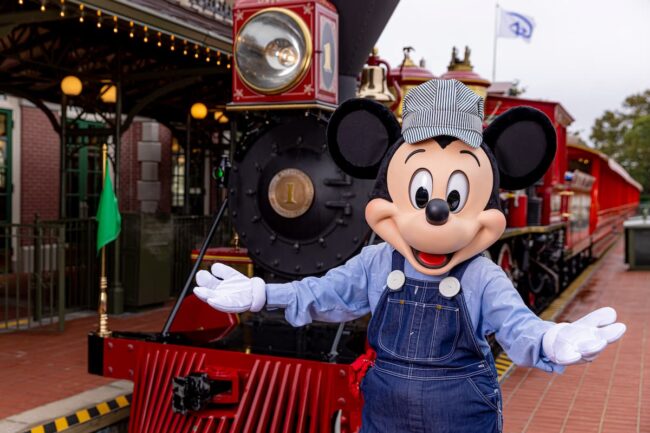 Mickey Mouse vestido de maquinista frente a un tren en Disney, invitando a los visitantes a vivir una experiencia mágica.