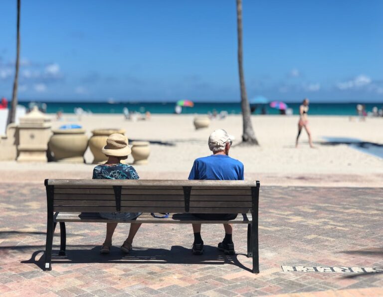 Pareja de turistas mayores sentados frente al mar en un día soleado, disfrutando de unas vacaciones relajadas en la playa.