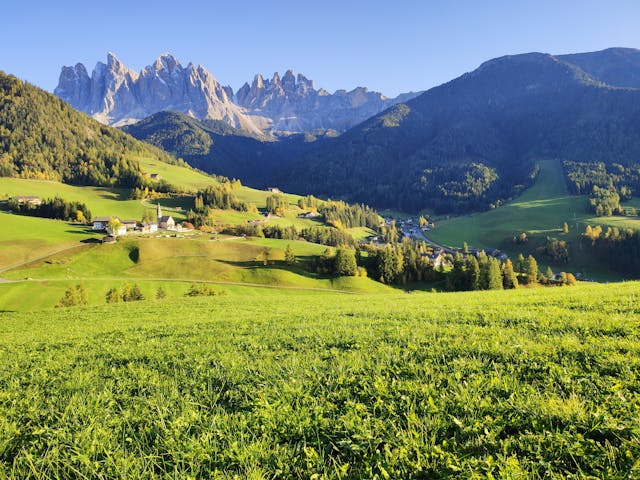 Paisaje alpino de los Dolomitas en Italia con montañas, praderas verdes y casas rurales.
