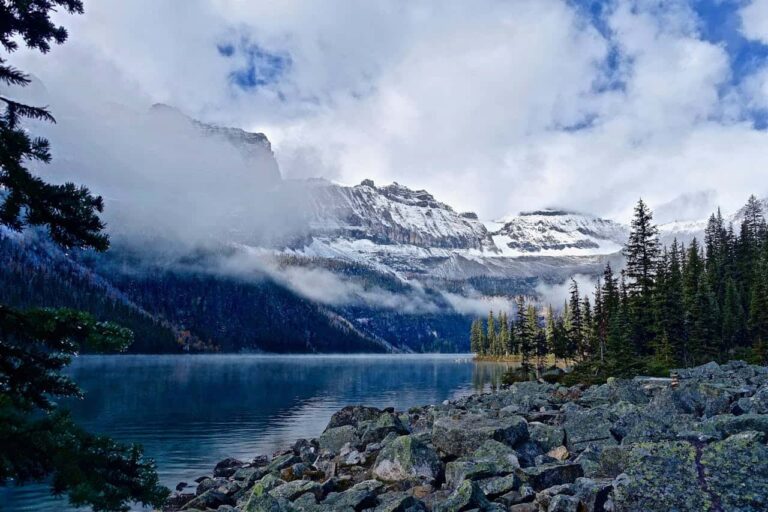 Lago rodeado de bosque y monta&ntilde;as nevadas en Alaska con nubes bajas sobre el paisaje