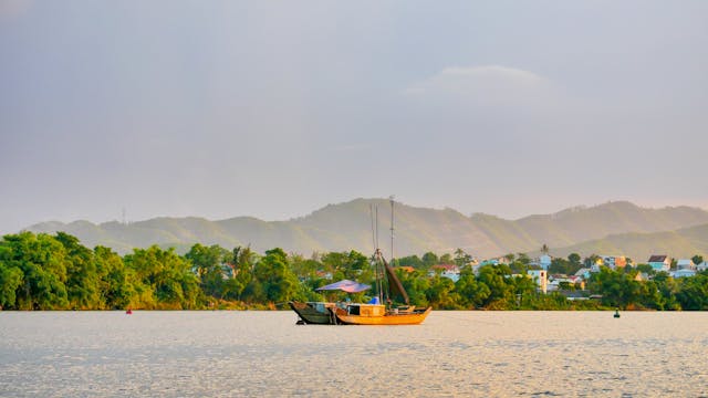Barca tradicional navegando por río de Vietnam al atardecer con montañas al fondo.