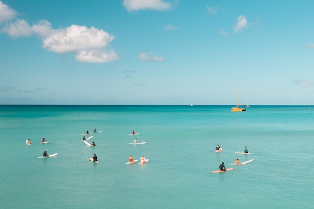 Personas practicando surf en aguas calmas y turquesas en Aruba con velero al fondo.