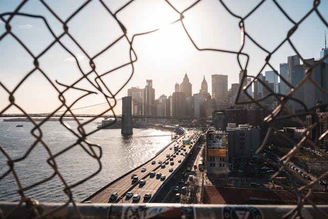 Vista de Manhattan desde un puente con reja en primer plano y tráfico en autopista junto al río.