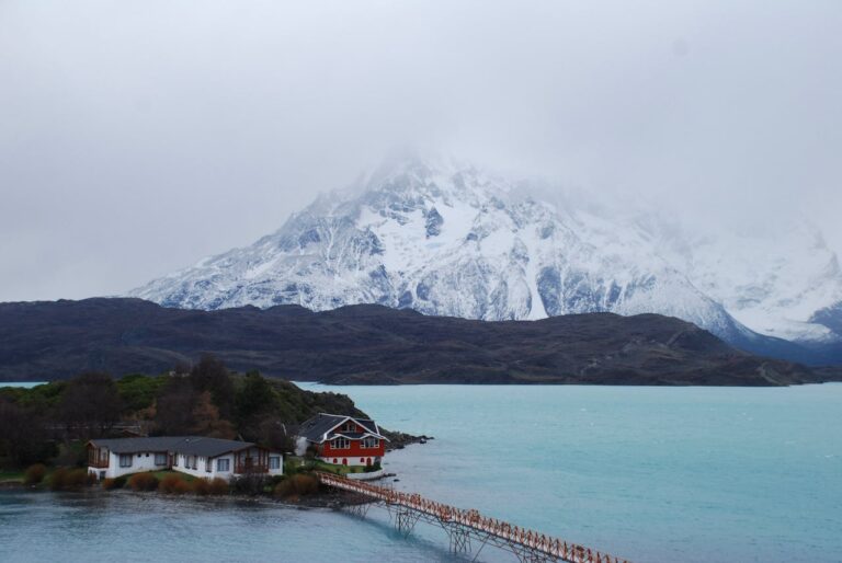Lago de color turquesa con monta&ntilde;as nevadas y caba&ntilde;as en la Patagonia chilena, una imagen que representa los paisajes ideales para unas vacaciones de invierno en Chile.