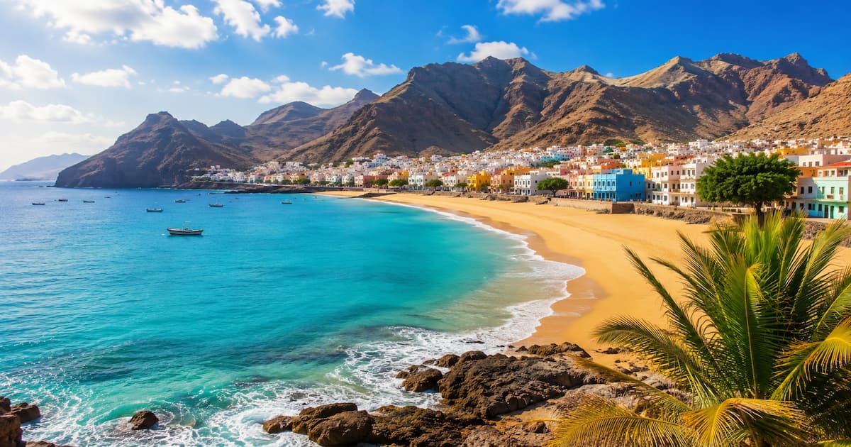 Vista panorámica de una playa de arena dorada en Cabo Verde, con mar turquesa, un pueblo costero de casas coloridas y montañas al fondo.