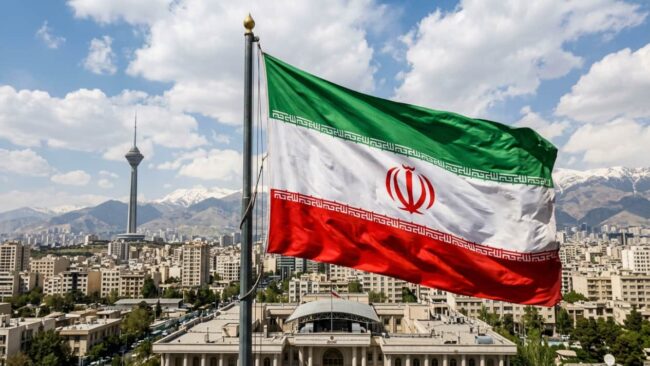 Bandera de Ir&aacute;n ondeando sobre Teher&aacute;n, capital del pa&iacute;s, con monta&ntilde;as al fondo y paisaje urbano.