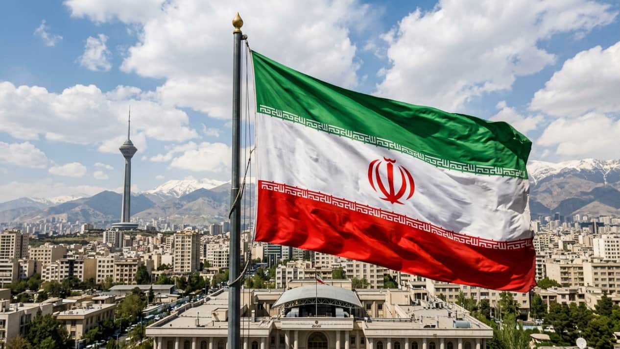 Bandera de Irán ondeando sobre Teherán, capital del país, con montañas al fondo y paisaje urbano.