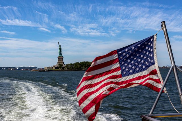 Bandera de Estados Unidos en un barco con la Estatua de la Libertad al fondo, imagen relacionada con dónde vivir el Fourth of July en Nueva York.