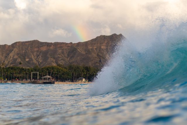 Ola en una playa de Hawaii con montañas al fondo y arcoíris sobre el océano Pacífico.