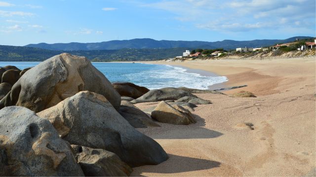 Playa extensa de arena dorada con rocas, mar azul y montañas al fondo, una imagen ideal para ilustrar playas largas de Europa.
