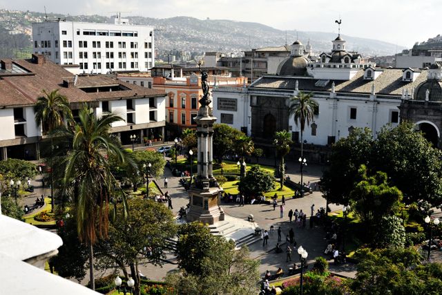 Vista de Quito con una plaza del centro histórico, monumento central, palmeras y edificios coloniales y modernos al fondo.
