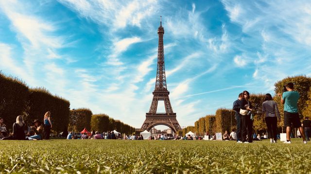 Alojarse cerca de la Torre Eiffel en París por primera vez Vista de la Torre Eiffel desde un parque de París, con turistas descansando sobre el césped y cielo despejado.