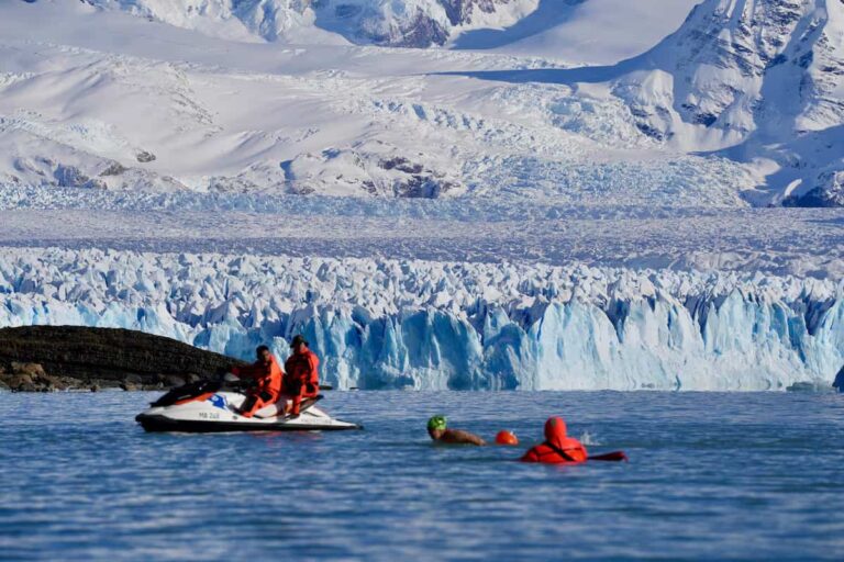 Nadador compitiendo frente al glaciar Perito Moreno con apoyo de seguridad en moto de agua durante una prueba en aguas heladas.