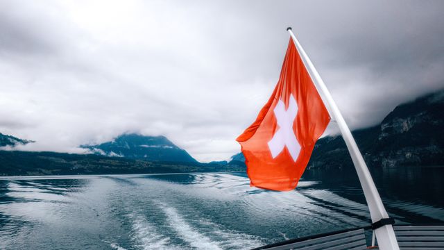Bandera de Suiza en los Alpes Bandera de Suiza en un barco sobre un lago rodeado de montañas, imagen relacionada con una guía sobre dónde queda Suiza.