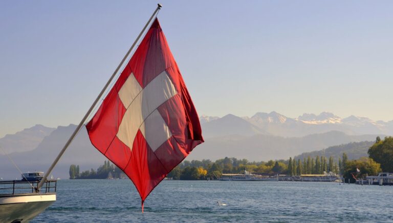 Bandera de Suiza en un barco sobre un lago, con monta&ntilde;as alpinas al fondo, imagen relacionada con d&oacute;nde queda Suiza.