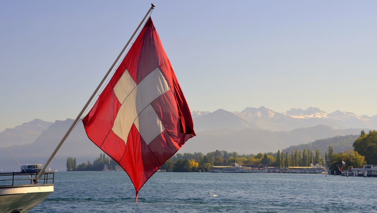 Bandera de Suiza en un barco sobre un lago, con montañas alpinas al fondo, imagen relacionada con dónde queda Suiza.