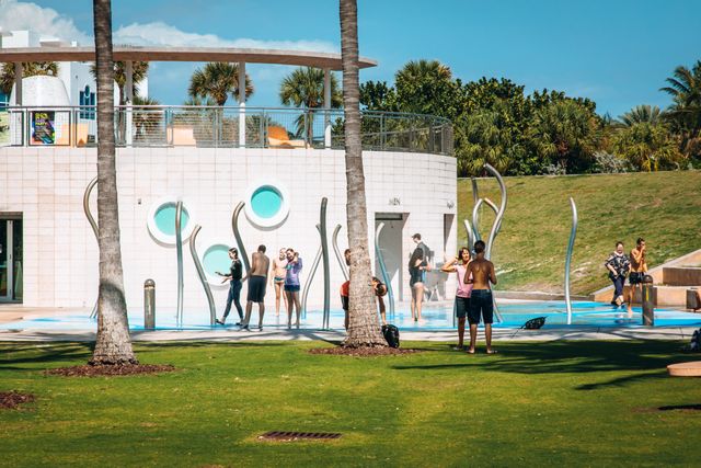 Niños y adultos disfrutando un área de juegos de agua en un parque de Miami, con palmeras y espacios verdes.