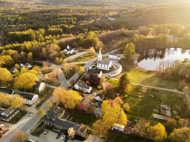 Vista aérea de un pequeño pueblo de Nueva Inglaterra en otoño, con árboles amarillos, casas, iglesia y paisaje rural.