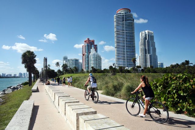 Personas andando en bicicleta por un paseo costero de Miami, con palmeras, edificios y vista al agua.