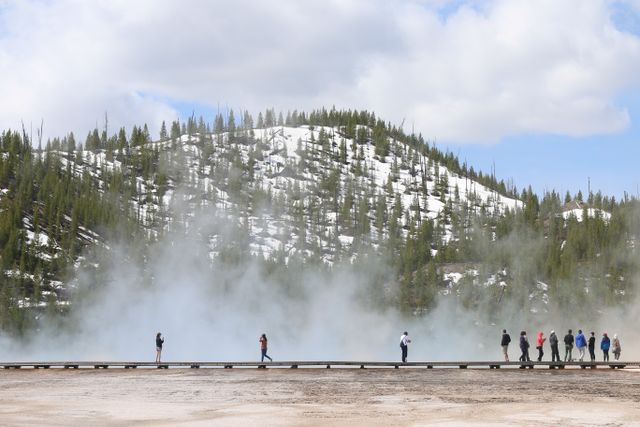 Visitantes caminando por una pasarela en una zona geotérmica de Yellowstone, con vapor, nieve, bosque y montañas al fondo.