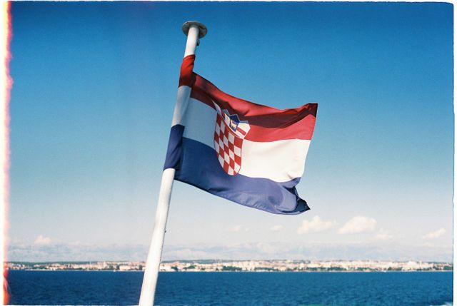 Mejor época para viajar a Croacia y recorrer su costa Bandera de Croacia ondeando frente al mar Adriático y una ciudad costera al fondo.