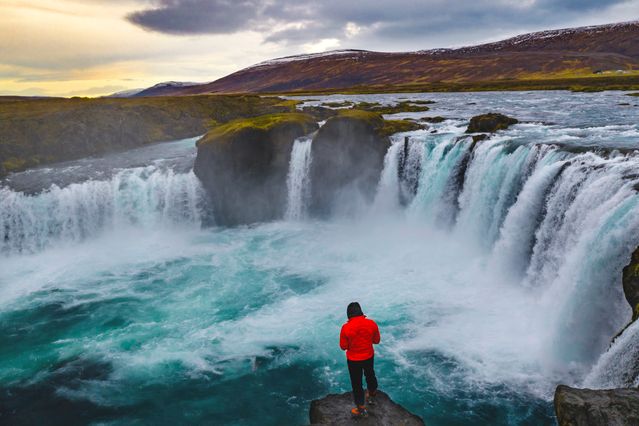Viajero frente a la cascada Goðafoss, rodeada de montañas y aguas turquesas en el norte de Islandia.