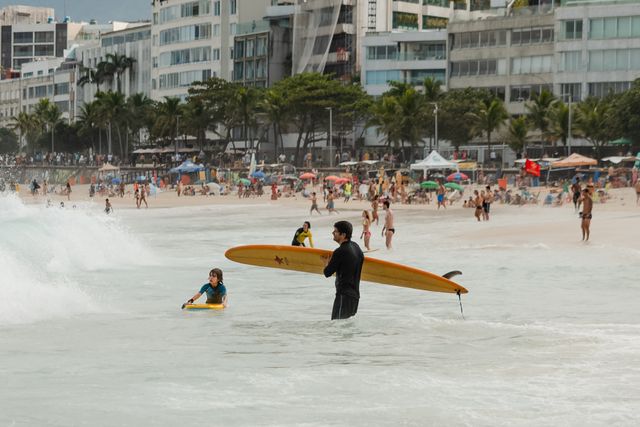 Adulto con tabla de surf y un niño jugando en el mar en una playa de Río de Janeiro, con edificios y palmeras al fondo.