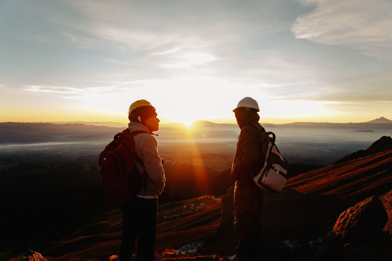Dos viajeros con mochilas contemplando el amanecer desde una zona de montaña