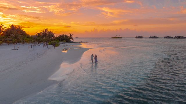 Atardecer en Maldivas durante la mejor época para viajar Pareja camina por una playa de Maldivas al atardecer con villas sobre el agua al fondo