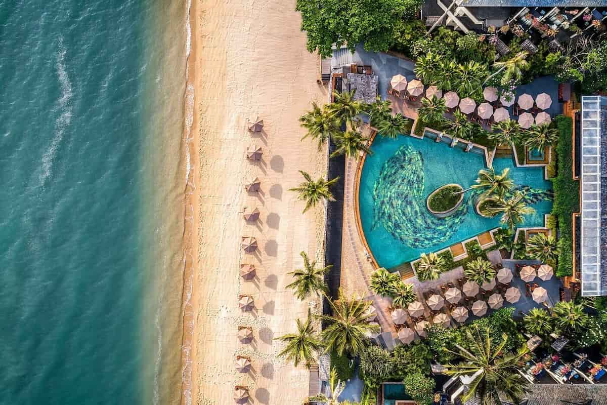 Vista aérea de un resort frente a la playa con piscina, palmeras y sombrillas junto al mar
